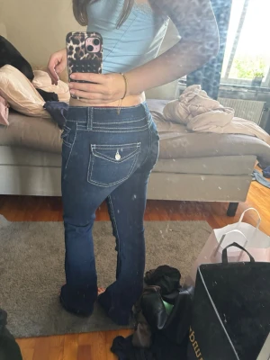 Mörkblå bootcut jeans med kontrastsömmar - Säljer ett par mörkblå bootcut jeans med vita kontrastsömmar och låg midja. Jeansen har fickor fram och bak, samt dekorativ knappdetalj på bakfickan. Klassisk denim-look som passar perfekt till crop tops och sneakers.