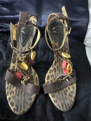 Bruna sandaler med leopardmönster Just Cavalli - Unika sandaler från Just Cavalli i brunt skinn med leopardmönstrad sula. Dekorerade med kedjor, berlocker och färgglada detaljer i guld, rött och träfärgat. Remmar runt ankeln och över foten ger en cool och trendig look.