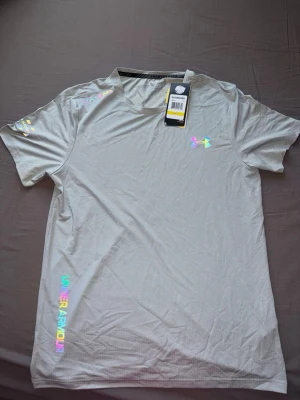 Ljusgrå t-shirt från Under Armour - Snygg ljusgrå t-shirt från Under Armour med holografiska loggor och textdetaljer på bröstet, ärmen och längs sidan. Tillverkad i ett mjukt och lätt syntetmaterial som andas, perfekt för träning eller chill. Klassisk passform med rund hals.