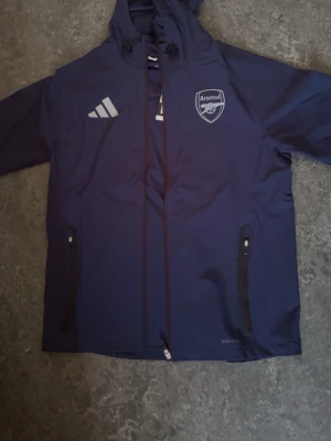 Mörkblå Arsenal vindjacka Adidas - Snygg mörkblå vindjacka från Adidas med Arsenal-logga på bröstet och vita Adidas-detaljer. Jackan har huva med dragsko, två dragkedjefickor och är tillverkad i lätt, vattenavvisande material. Perfekt för dig som vill visa ditt stöd för Arsenal.