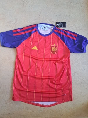 Spanien fotbollströja - Säljer en officiell matchtröja från Spaniens landslag i rött med lila ärmar och röda detaljer. Tröjan har gula ränder och landslagets emblem på bröstet. Tillverkad i lätt och ventilerande funktionsmaterial, perfekt för fotboll eller träning. Ny med prislappar.