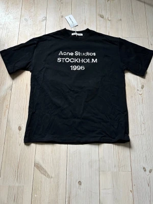 Svart Acne Studios t-shirt Stockholm - Svart t-shirt från Acne Studios med vit text 'STOCKHOLM 1996' framtill. Klassisk passform och rund hals. Perfekt för dig som gillar stilrena och trendiga plagg med en urban vibe. Sitter bäst på folk med S. Fraktar samma dag nästan alltid! Ställ frågor ifall osäker på nått!