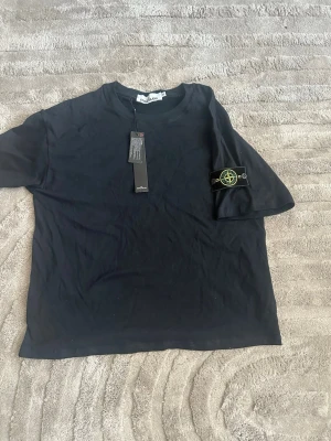 Svart t-shirt från Stone Island - Snygg svart t-shirt från Stone Island med klassisk logga på vänster ärm. T-shirten har rund hals och korta ärmar, tillverkad i mjuk bomull för en skön känsla. Perfekt för dig som gillar streetwear och vill ha en stilren look.