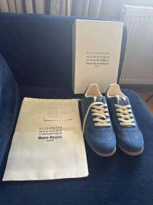 Blå Maison Margiela sneakers - Maison Margiela sneakers i blå mocka och läder med vita detaljer och vita skosnören. Klassisk låg modell med beige innersula och gummisula. Snygg och stilren design som passar till många outfits.