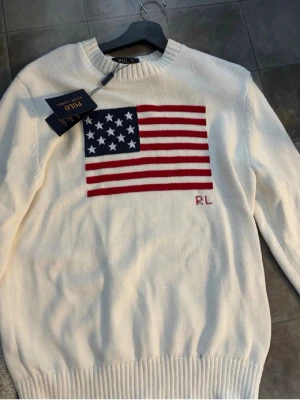 Vit stickad tröja Polo Ralph Lauren - Äkta och oanvänd vit stickad tröja från Polo Ralph Lauren med amerikansk flagga i rött, vitt och blått på bröstet samt RL-broderi. Tröjan har rund halsringning och ribbade muddar vid ärmslut och nederkant. Perfekt för dig som gillar klassisk och preppy stil.