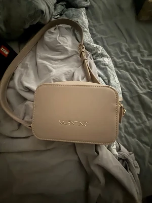 Beige handväska från Mario Valentino - Snygg beige handväska från Mario Valentino i stilren, rektangulär modell. Väskan har justerbar axelrem och guldfärgade dragkedjor. Tillverkad i slätt konstläder med diskret logga framtill. Perfekt för dig som gillar minimalistisk design.