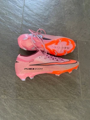 Rosa Nike Air Zoom fotbollsskor - Säljer ett par rosa Nike Air Zoom elite fotbollsskor med svart swoosh och textdetaljer. Skorna har en låg profil, snörning och en yttersula i plast med orange och genomskinliga dobbar för bra grepp på planen. Perfekta för dig som vill sticka ut på matchen!