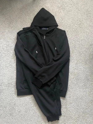 Svart Polo Ralph Lauren tracksuit - Säljer en svart hoodie från Polo Ralph Lauren med dragkedja framtill och klassisk huva. Tröjan har det ikoniska vita Polo-logotrycket på bröstet och ribbade muddar vid ärmar och nederkant. Perfekt för en chill och stilren look.
