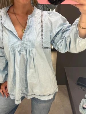 Ljusblå blus från Molin Copenhagen - Ljusblå blus med v-ringning och dekorativa broderidetaljer från Molin Copenhagen. Blusen har långärmade puffärmar och veckade detaljer framtill. Tillverkad i ett tunt, luftigt material som ger en avslappnad och trendig look.