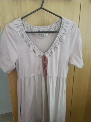 Ljusrosa kort klänning med snörning - Söt ljusrosa klänning med kort ärm och v-ringning. Klänningen har rynkad kant vid halsen och en dekorativ snörning framtill. Modellen är kort och har en avslappnad passform, perfekt för varmare dagar.