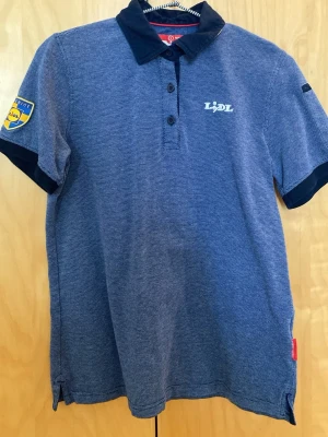 Blå piké t-shirt med Lidl-logga - Säljer en blå piké t-shirt med Lidl-logga på bröstet och ett Lidl-märke på ärmen. Tröjan har mörkblå krage och ärmslut samt knappar vid halsen. Perfekt för dig som gillar unika plagg med lite humor.