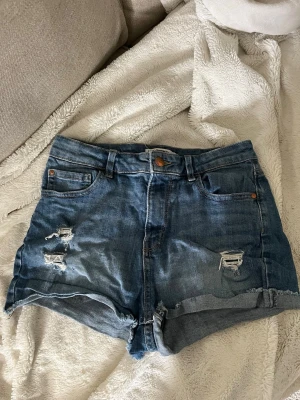Blå denimshorts med slitningar - Säljer ett par blå denimshorts från Detroit med råa kanter och slitna detaljer framtill. Shortsen har fem fickor, bälteshällor och knappgylf. Perfekta för sommaren och ger en avslappnad vibe.