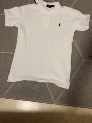 Vit pikétröja från Polo Ralph Lauren - Klassisk vit pikétröja från Polo Ralph Lauren med blå logga broderad på bröstet. Tröjan har korta ärmar, krage och knappar framtill. Perfekt för en clean och stilren look.
