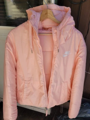 Vändbar Ljusrosa pufferjacka från Nike - Säljer en ljusrosa pufferjacka från Nike med huva och dragkedja. Jackan har två sidofickor, justerbar huva med snören och stor vit Nike-logga på bröstet samt insidan. Perfekt för kalla vinterdagar och riktigt snygg streetstil.Vändbar 