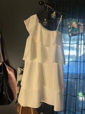 Vit oneshoulder klänning Gina Tricot - Säljer en vit oneshoulder klänning från Gina Tricot med volangdetaljer och asymmetrisk axel. Klänningen har ett lager-på-lager utseende och är perfekt för sommarens alla tillfällen. Tillverkad i ett lätt och luftigt material.