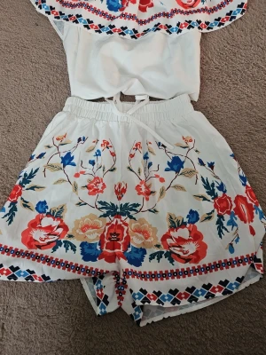 Vitt set med blommigt mönster - Säljer ett superfint tvådelat set i vitt med färgglada blommor i rött, blått och gult. Toppen är off shoulder med volang och resår, och shortsen har hög midja och elastisk linning. Perfekt för sommardagar och festivaler!
