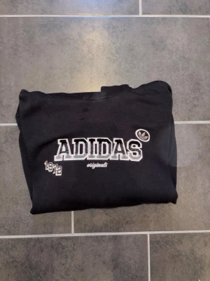 Svart hoodie från Adidas Originals - Svart hoodie från Adidas Originals med vit broderad logga och texten 'ADIDAS 1972 originals' på bröstet. Klassisk huva och känguruficka framtill. Perfekt för en avslappnad och sportig stil.