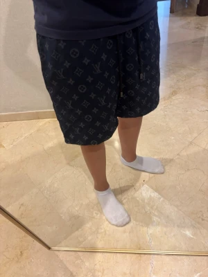 Mörkblå shorts med monogrammönster - Säljer ett par mörkblå shorts med ikoniskt monogrammönster och snörning i midjan. Shortsen har en avslappnad passform och elastisk midja för extra komfort. Perfekta för sommardagar eller när du vill ha en chill look.