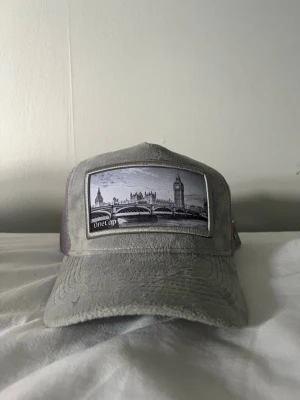 Grå keps med Londonmotiv Onecap - Snygg grå keps från Onecap med mesh på sidorna och baksidan. Framtill finns ett broderat motiv av London med Big Ben och Westminster. Justerbar snapback och böjd skärm. Perfekt för dig som gillar streetstyle och unika detaljer.
