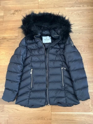 Svart dunjacka med fuskpäls - Snygg svart dunjacka med huva och avtagbar fuskpäls. Jackan har två dragkedjeförsedda fickor framtill och quiltad design. Perfekt för kalla vinterdagar och ger en stilren look. Märke syns ej tydligt på bilden.
