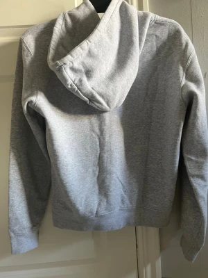 Grå hoodie med tryck från Bvaldi  - Snygg grå hoodie från Bvaldi med svart tryck framtill och känguruficka. Klassisk modell med huva och dragsnören. Tillverkad i mjukt sweatshirtmaterial, perfekt för en avslappnad stil.