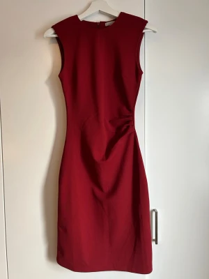 Vinröd ärmlös klänning från H&M - Snygg vinröd klänning från H&M i figurnära modell. Klänningen är ärmlös, har rund halsringning och en diskret drapering i midjan. Tillverkad i ett mjukt och stretchigt material som ger en bekväm passform.