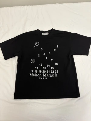 Maison Margiela svart t-shirt - Svart t-shirt från Maison Margiela med ikoniskt tryck av siffror och logga på framsidan. Klassisk passform med rund hals och korta ärmar. Tillverkad i mjuk bomull som känns skön mot huden. Perfekt för dig som gillar stilrena och trendiga plagg.