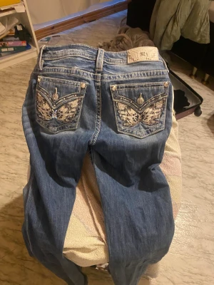 Blå jeans med broderade fickor - Säljer ett par blåa miss me jeans med snygga broderade detaljer och nitar på bakfickorna. Jeansen har kontrastsömmar, fem fickor och normal midja. Perfekta för dig som vill sticka ut lite extra med coola detaljer på jeansen.