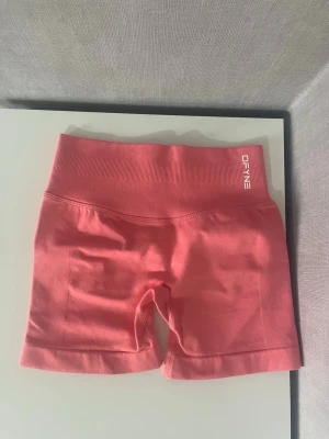 Rosa träningstights shorts från DFYNE - Säljer ett par rosa träningstights shorts från DFYNE med hög midja och ribbad linning. De är tillverkade i ett stretchigt material som passar perfekt för gymmet eller andra träningspass. Snygg och bekväm passform som framhäver formerna.