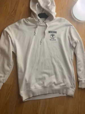 Vit Moschino hoodie - Gör dig redo för hösten redan nu!  Jag säljer nu min stilrena Moschino hoodie med luva (vit). Står ingen storlek men skulle vara rätt säker om jag sa L.  Anledning? Aldrig använd. För dyr för o bara hänga i garderoben.  Skick? Mycket bra. Inte använt mycket alls, bara hängt i garderoben.  Pris? 600 men priset går självklart att diskuteras med rimlig mängd!  Frågor eller nyfiken? Hör av dig!