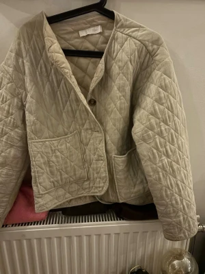 Beige quiltad cardiganjacka - Quiltad beige cardiganjacka med stora framfickor och knäppning med tre knappar. Jackan har en boxig passform och är tillverkad i ett mjukt, vadderat material med rutmönster. Perfekt för lager-på-lager och en chill stil.