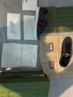 Gucci sneakers med monogram och röd-blå rand - Snygga Gucci sneakers i lågt snitt med mörkgrått monogrammönster och klassiska röd-blå ränder på sidan. Skorna har svart snörning och vit sula. Materialet är canvas med läderdetaljer. Perfekta för dig som vill ha en exklusiv och trendig look.