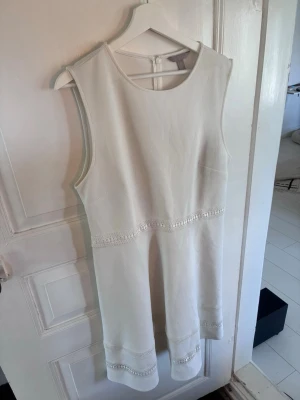 Vit ärmlös klänning med spetsdetaljer - Snygg vit ärmlös klänning med rund halsringning och dragkedja i ryggen. Klänningen har fina spetsdetaljer vid midjan och nederkanten som ger en elegant touch. Perfekt för sommarens alla tillfällen.