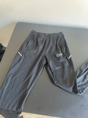 Svarta träningsbyxor från Nike - Säljer ett par svarta träningsbyxor från Nike med vit logga på vänster ben och diskreta vita detaljer vid fickorna. Byxorna har resår i midjan och är tillverkade i ett lätt och bekvämt material som passar perfekt för träning eller chill.