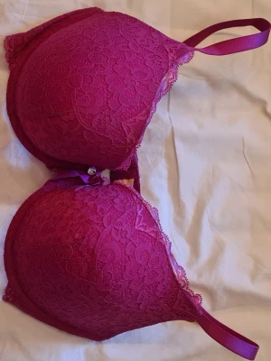 Mörkrosa hunkemöller push-up bh med spets - Snygg mörkrosa push-up bh från hunkemöller med vadderade kupor och spetsdetaljer. Bh:n har den lilla rosetten och metallberlocken framtill mellan kuporna. Axelbanden är justerbara och materialet är en kombination av spets och slät textil.     Den är i storlek 85E - men den passar även mig som har 80C. Super bra skick, endast använd en gång!