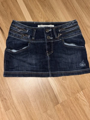 Mörkblå jeanskjol med dragkedjor - Snygg mörkblå jeanskjol i kort modell med dubbla knappar framtill, dekorativa dragkedjor och fickor både fram och bak. Tuffa detaljer och kontrastsömmar ger kjolen en cool look. Perfekt till sneakers eller boots.