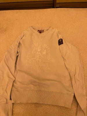Vit fleece tröja med tryck Parajumpers - Säljer en vit fleece tröja i storlek M med tryck framtill och patch på ena ärmen. Tröjan har rund halsringning och ribbade muddar vid ärmslut och nederkant. Perfekt för dig som gillar en enkel men snygg stil.