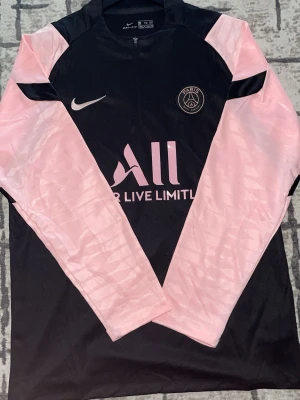 PSG träningströja  Nike - Snygg långärmad träningströja från PSG i svart med rosa ärmar och detaljer. Tillverkad i lätt funktionsmaterial, perfekt för fotbollsträning. Står XL men passar skön som L. Dropa vikten och längden så svarar jag ifall den passar.