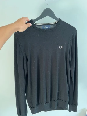 Svart stickad tröja från Fred Perry - Klassisk svart stickad tröja från Fred Perry med rund halsringning och diskret logga broderad på bröstet. Tröjan har långa ärmar och ribbade muddar vid ärmslut och nederkant. Perfekt för en stilren och enkel look.