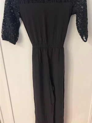 Svart jumpsuit med spetsärmar och knyt - Snygg svart jumpsuit med halvlånga spetsärmar och elastisk midja. Baksidan har ett knytband i midjan för extra passform. Perfekt för dig som vill ha en stilren och trendig look med fina spetsdetaljer på ärmarna.Storlek 10-12år 