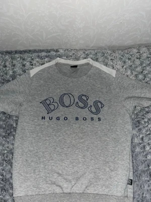 Grå kortärmad topp från Hugo Boss - Snygg grå kortärmad topp från Hugo Boss med blå logga framtill och vita detaljer på axlarna. Tillverkad i mjukt bomullsmaterial och har en avslappnad passform. Perfekt för en stilren och enkel look.