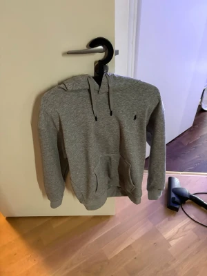 Grå hoodie med ficka och huva - Säljer en stilren grå hoodie med huva och dragsnören. Hoodien har en klassisk känguruficka framtill och ribbade muddar vid ärmar och midja. Perfekt för chill dagar eller när du vill ha en enkel och snygg look. Säljer pga för liten.