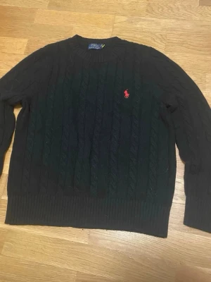 Mörkgrön kabelstickad tröja Polo Ralph Lauren - Kabelstickad tröja i mörkgrönt från Polo Ralph Lauren med klassisk röd logga broderad på bröstet. Tröjan har rund halsringning, långa ärmar och ribbade muddar. Perfekt för dig som gillar tidlös och stilren design.
