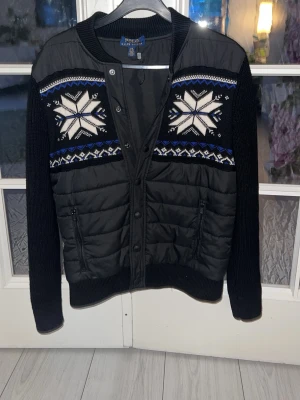Ralph Lauren cardigan - Svart quiltad cardiganjacka från Polo Ralph Lauren med stickade ärmar och ribbade muddar. Framsidan och baksidan har vita och blå snöstjärnemönster samt dragkedjefickor. Jackan har tryckknappar och en sportig, vinterinspirerad look.