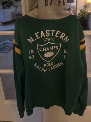 Grön hoodie från Polo Ralph Lauren - Grön hoodie från Polo Ralph Lauren med stora RL-bokstäver och broderad logga framtill. Har gula och mörkblå ränder på ärmarna, grå huva och känguruficka. Tryck och brodyr på både fram- och baksida. Perfekt för en sportig och avslappnad stil.
