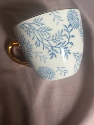 Vit kopp med blått blommönster - Säljer en vit porslinskopp med blått blommönster och guldfärgat handtag. Koppen har en klassisk form och det detaljerade mönstret ger en elegant känsla. Perfekt för te eller kaffe och blir en fin detalj i köket.