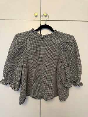 Rutig blus med puffärm och volang - Snygg svartvit rutig blus med puffiga trekvartsärmar och volangdetaljer vid hals och ärmslut. Blusen har en rak passform och dragkedja i ryggen. Perfekt för dig som gillar en trendig och söt stil.