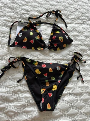 Svart bikini med fruktmönster och knyt - Snygg svart bikini med färgglatt fruktmönster, bland annat vattenmelon, kiwi och ananas. Både överdel och underdel har knytband i sidorna och nacken för justerbar passform. Perfekt för lata dagar på stranden eller poolen. Super fin och super somrig storlek s och helt ny med prislappar säljer då jag köpte fel storlek annars helt perfekt!🏝️❤️🥹