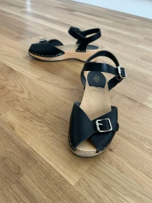 Fina  sandaler från Hasbeens  - Snygga svarta sandaler med träbotten och klassisk design. Remmar i svart läder med silverfärgade spännen över foten och runt ankeln. Sulan är i ljust trä och har en liten klack som är 5 cm.. Perfekta för dig som gillar retrovibbar och stilren look.