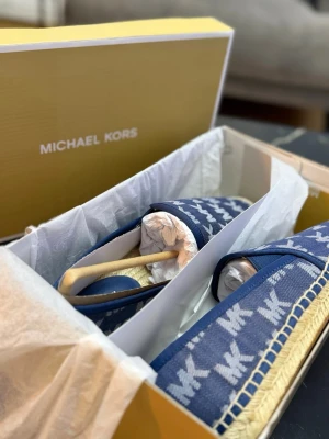 Blå ballerinaskor från Michael Kors - Snygga blå ballerinaskor från Michael Kors med ikoniskt MK-mönster. Ovandelen är i textil och sulan i gummi. Skorna har en flätad detalj runt sulkanten och en klassisk, rundad tå. Perfekta för dig som gillar stilrena och trendiga skor.